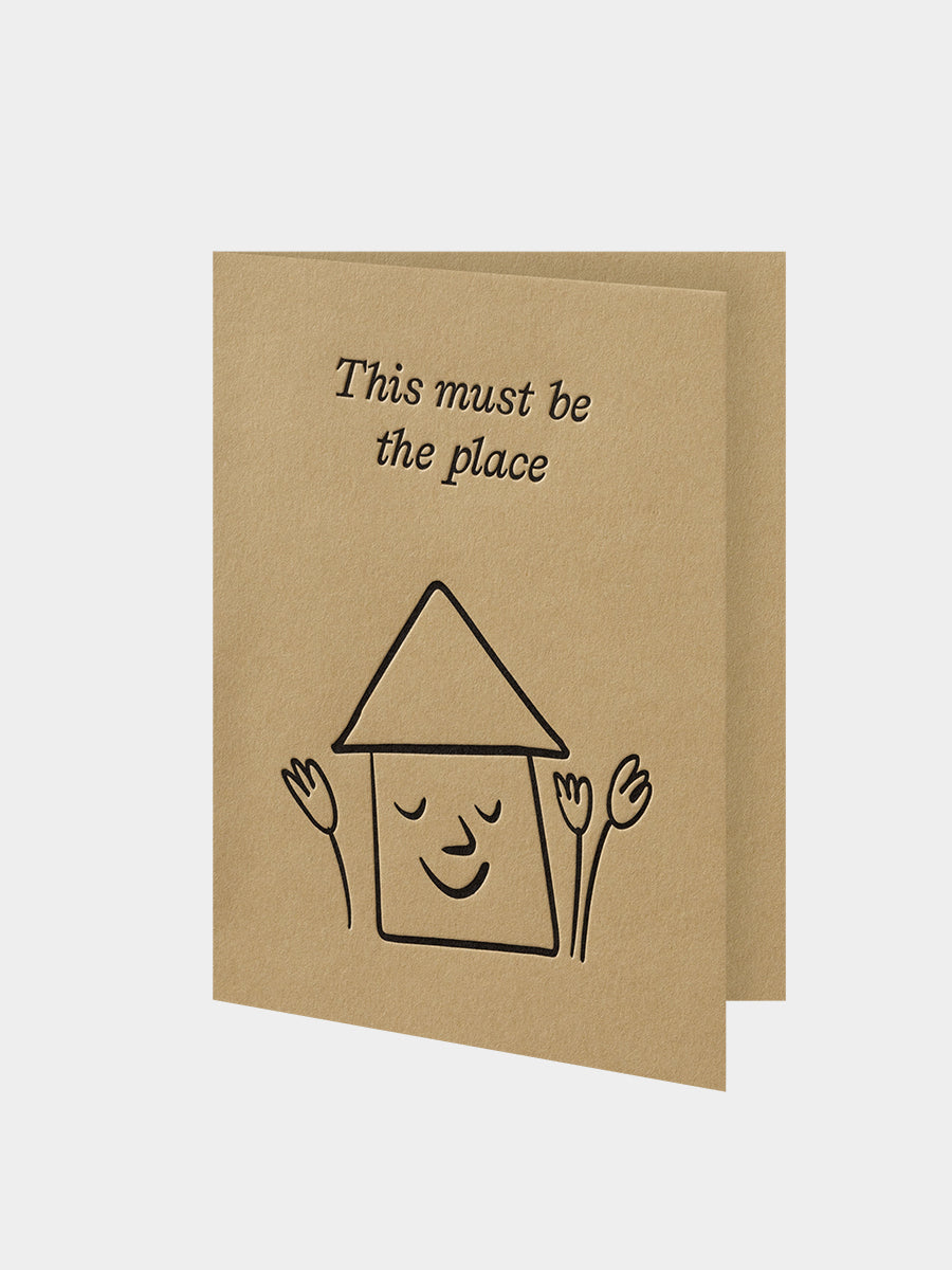 Kraft paper card with a house illustration and text 'This must be the place' on a light gray background